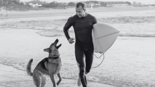 VERILOX® | The image shows a surfer in a wetsuit walking along the beach. He is carrying a surfboard under his arm. A dog is running beside him, jumping up happily. The sea is visible in the background.