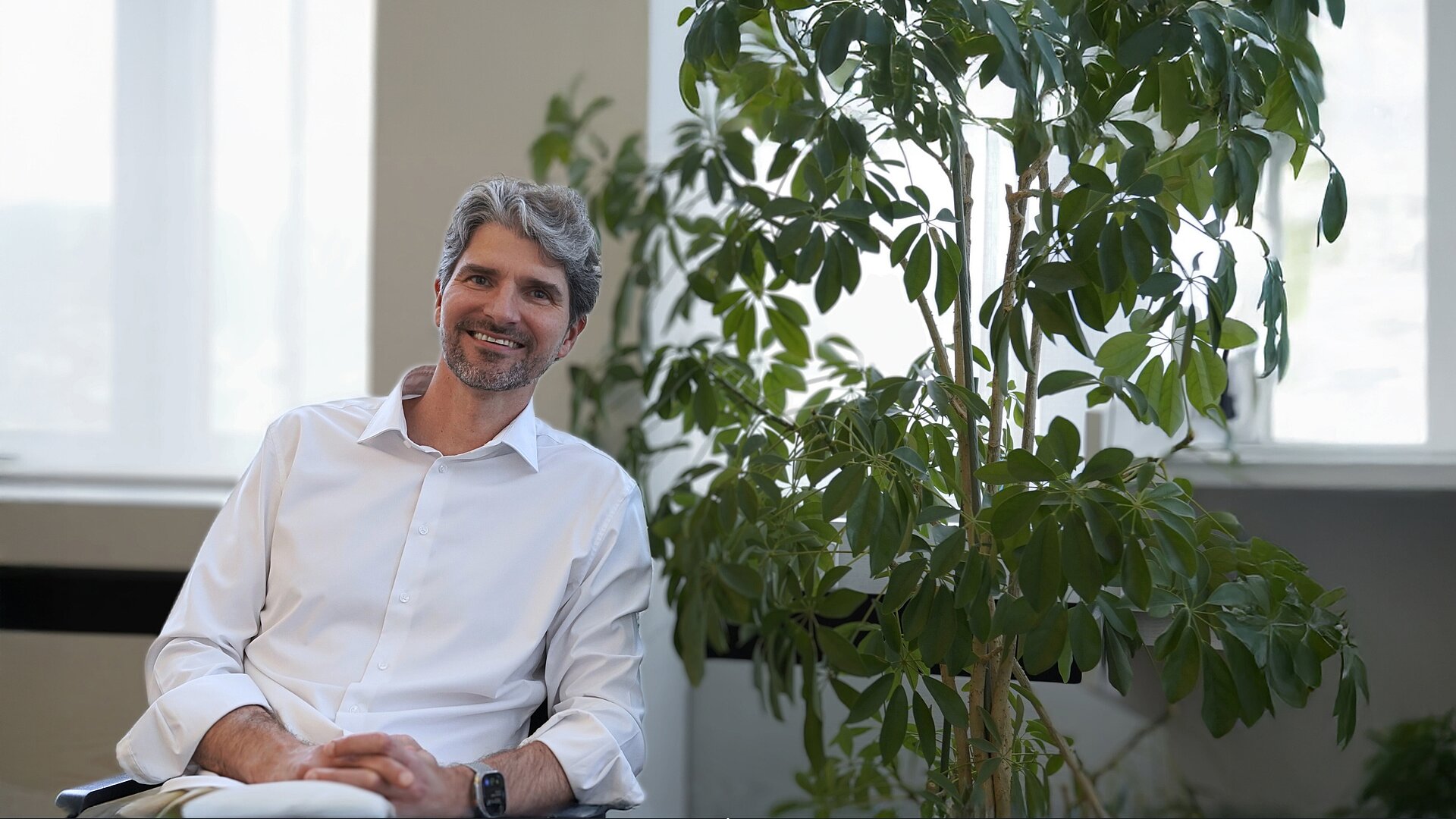 Dr. Alessandro Alan Porporati, wearing a white shirt, sits in a modern office next to a large indoor plant. He is giving an interview on hip arthroplasty, clinical evidence in orthopedics, and high-performance ceramics in dental implantology.